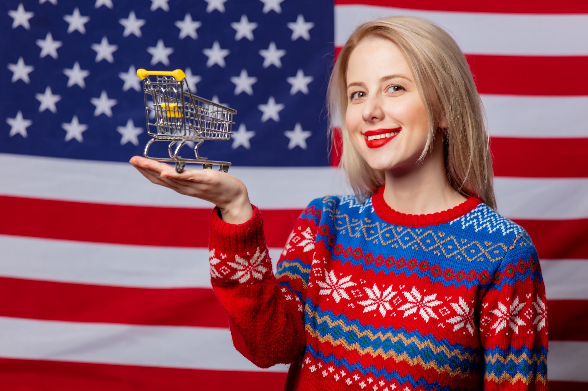 Piękna blondynka w świątecznym swetrze trzyma koszyk na fladze USA