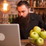 Przystojny barman sprawdza zamówienia na swoim laptopie. Hipsterski pub.
