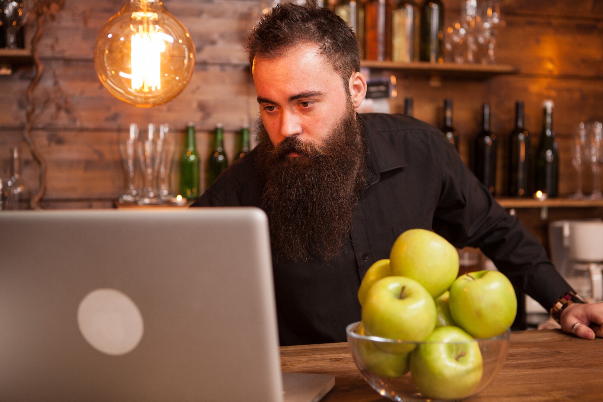 Przystojny barman sprawdza zamówienia na swoim laptopie. Hipsterski pub.