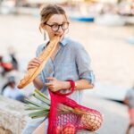 Styl życia portret młodej kobiety z siatkową torbą pełną świeżej żywności jedzącej bagietki na świeżym powietrzu na starym mieście