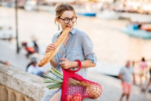 Styl życia portret młodej kobiety z siatkową torbą pełną świeżej żywności jedzącej bagietki na świeżym powietrzu na starym mieście