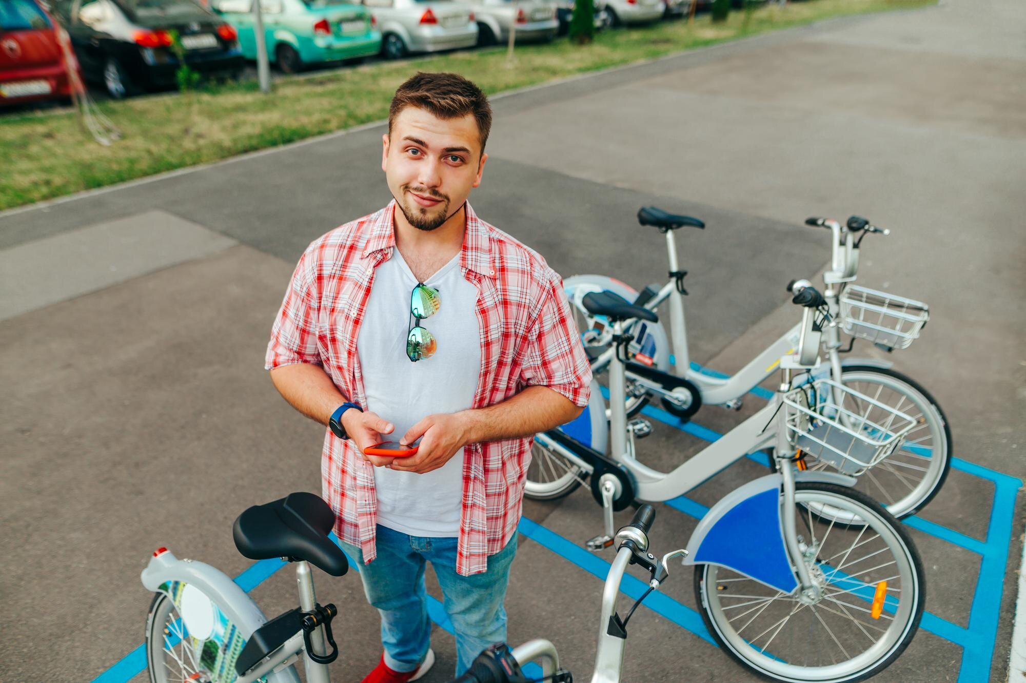 Pozytywny młody człowiek stojący w rzędzie, dzielący rower ze smartfonem w dłoni, patrzący w kamerę i uśmiechający się Wesoły mężczyzna bierze rower dzielący na spacer online