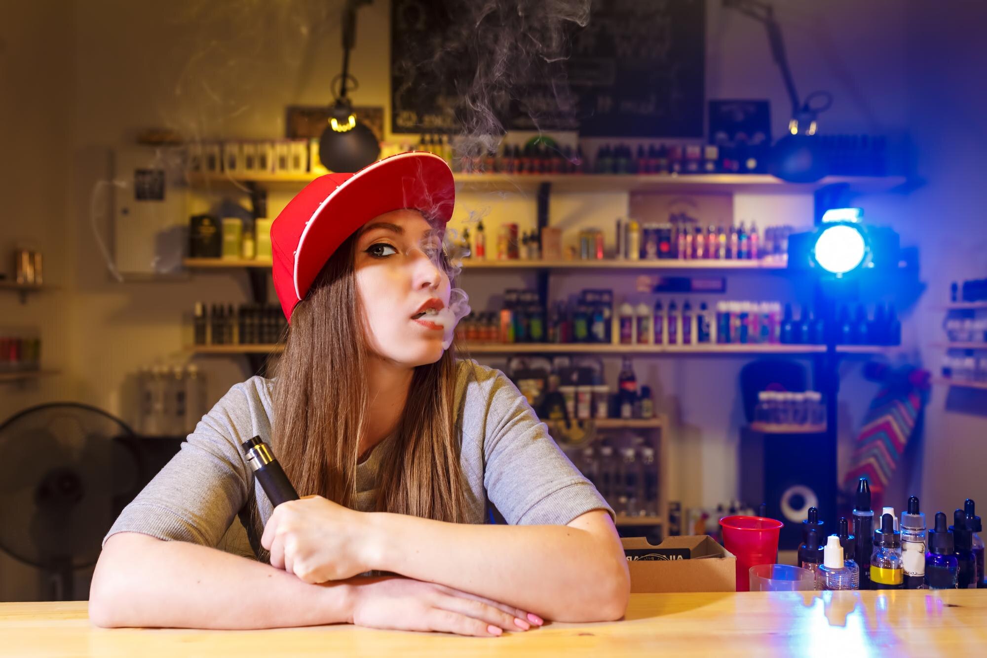 Młoda ładna kobieta w czerwonej czapce pali elektroniczny papieros w sklepie vape