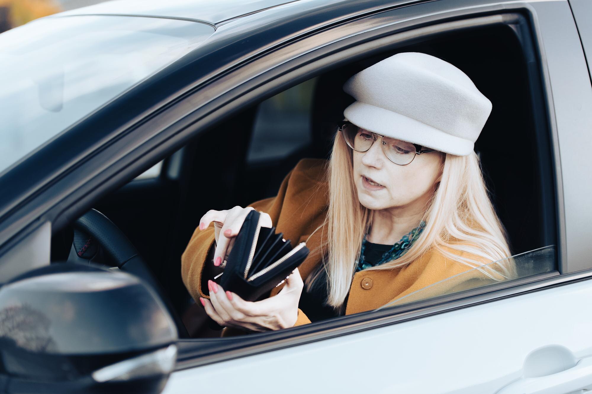 Kobieta w średnim wieku siedzi za kierownicą samochodu i szuka pieniędzy w portfelu, aby zapłacić za parking
