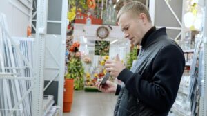 Mężczyzna uważnie czyta opakowania produktów w sklepie z narzędziami, otoczony półkami z artykułami do poprawy domu