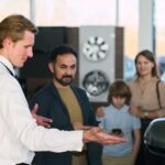 Young adult man showing car to multiethnic family at dealership
