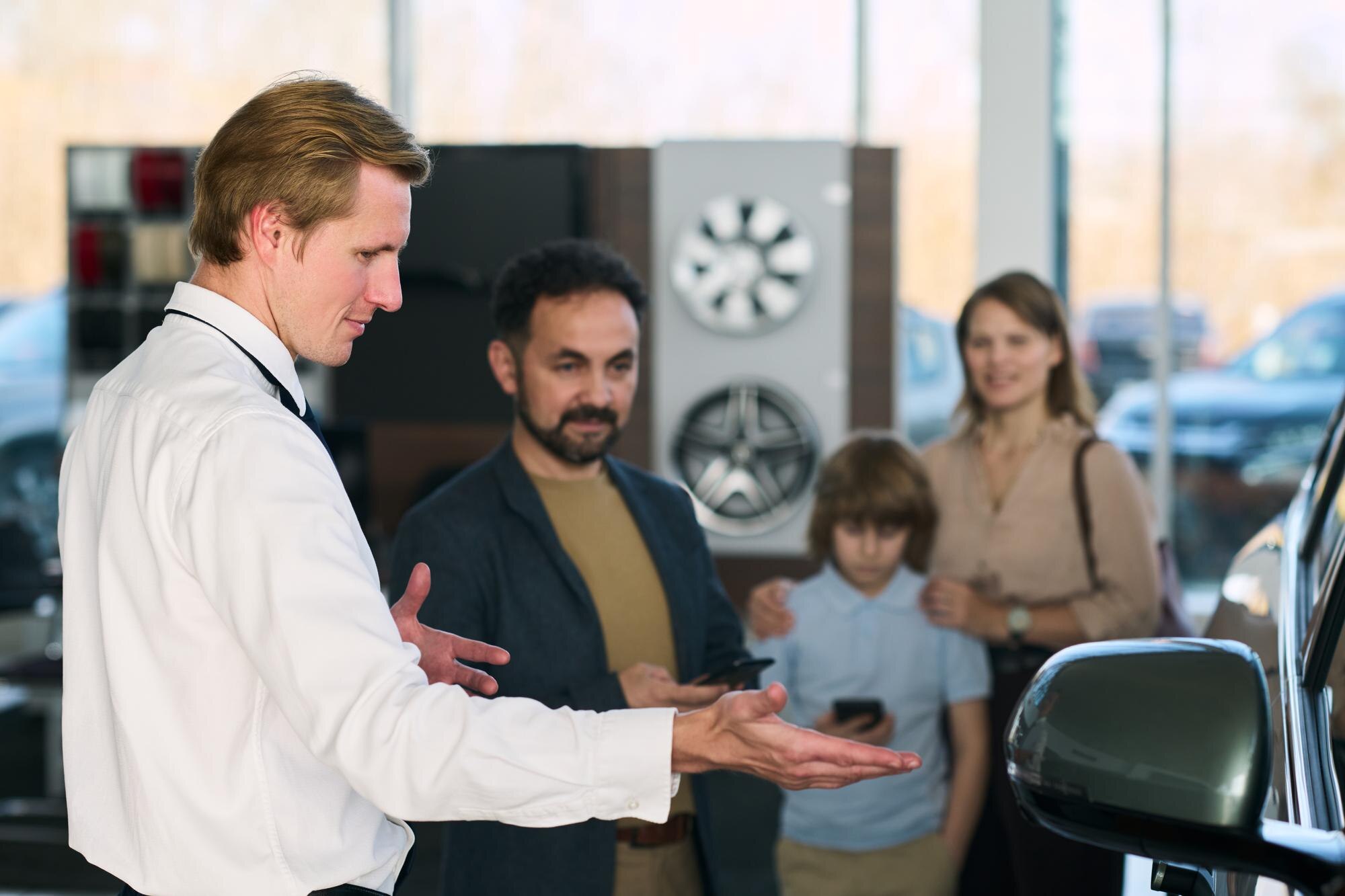 Young adult man showing car to multiethnic family at dealership