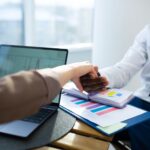 Two business professionals shaking hands over graphs and reports in an office setting.
